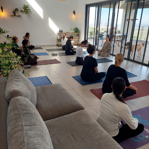 מתאמנות עושות יוגה בסלון של בית אשר המתאמנות יושבות על מזרן שחור כחול ואדום והמדריכה יושבת על הרצפה