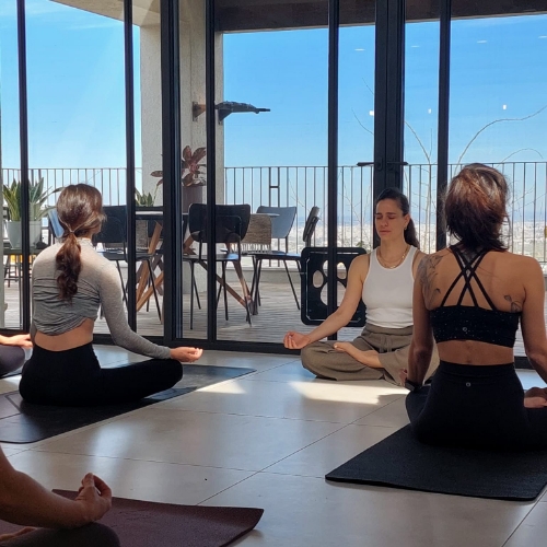 מתאמנות עושות יוגה בסלון של בית אשר המתאמנות יושבות על מזרן שחור כחול והמדריכה יושבת על הרצפה