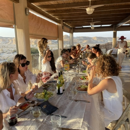 קבוצה גדולה של נשים אשר כולן לבושות בלבן יושבות ליד שולחן אוכל ארוך מול נוף של הרים ועל השולחן שלל של ארוחות ושתייה