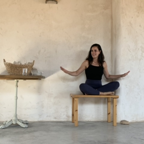 אישה צעירה משלבת רגליים יושבת על כיסא מעץ ומרימה ידיים ולידה שולחן עץ ועליו סלסלה מקש